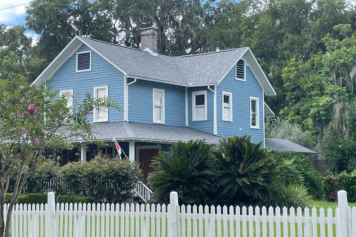 A longtime family home in Mount Dora Blue two-story farmhouse-style home with a wraparound porch behind a white picket fence, the kind of longtime family home people leave when downsizing in Mount Dora