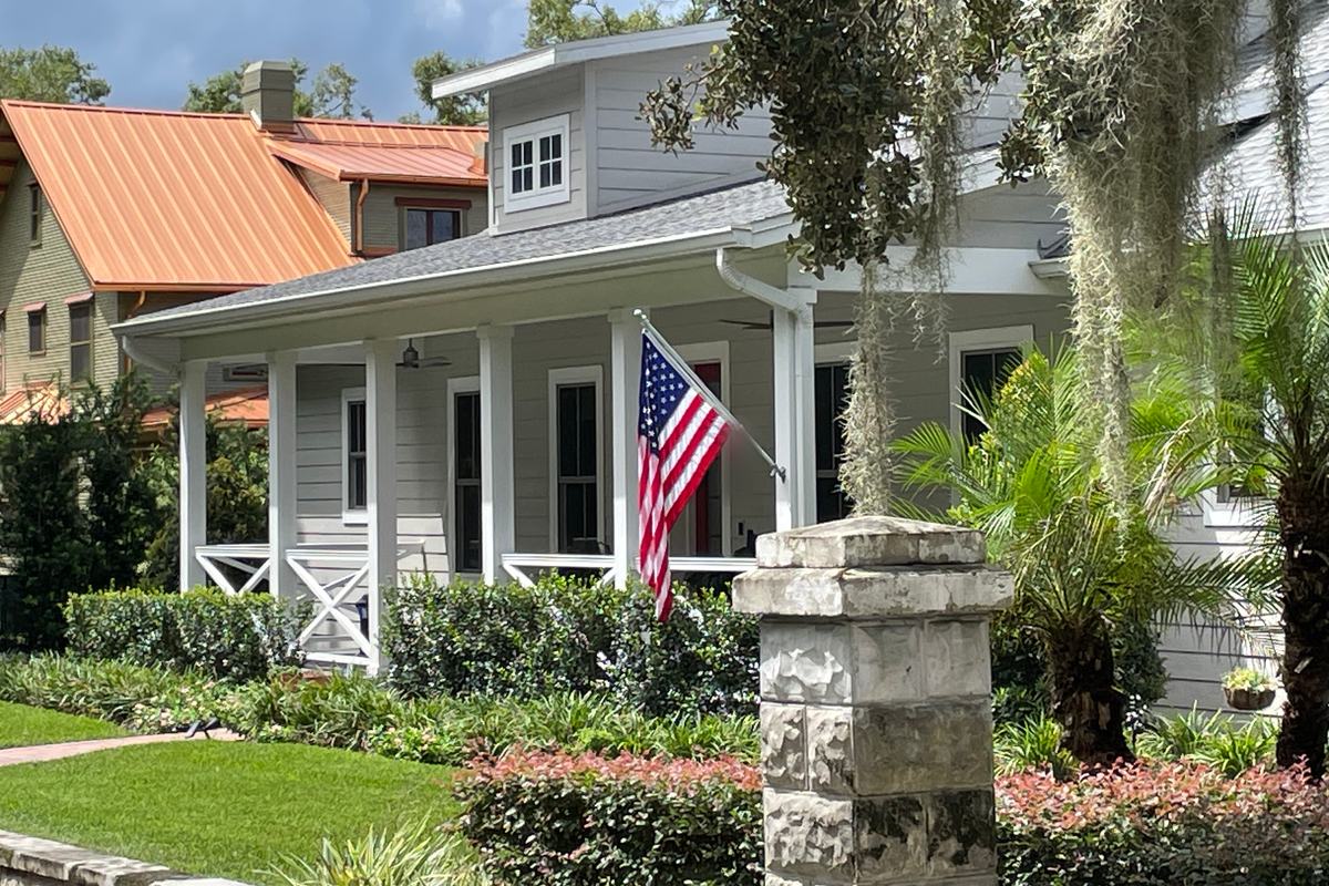 Gray older home in Mount Dora with a white-columned front porch, an American flag, and a stone pillar: curb appeal worth weighing when prepping an older home to sell