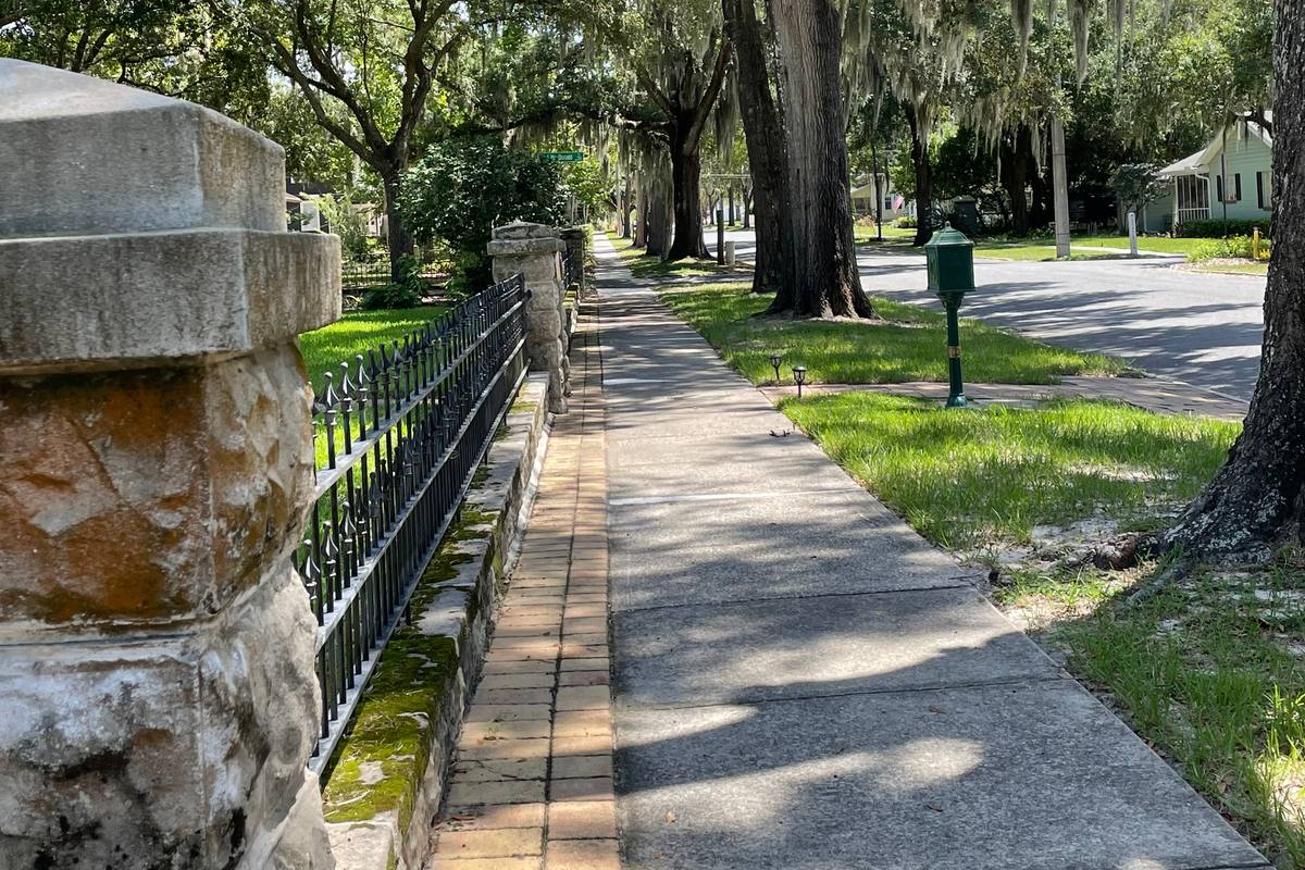 Shaded sidewalk, a rough-stone pillar, and an iron fence under moss-draped oaks on a street of older Mount Dora homes